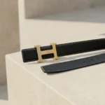 Premium womens black grained leather belt with a brushed gold H shaped buckle on a minimalist stone pedestal (6)